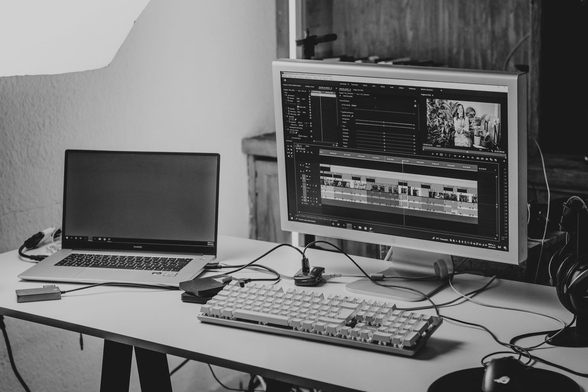 Black-and-white photo of a desk with a laptop, a large monitor showing video editing software and a keyboard