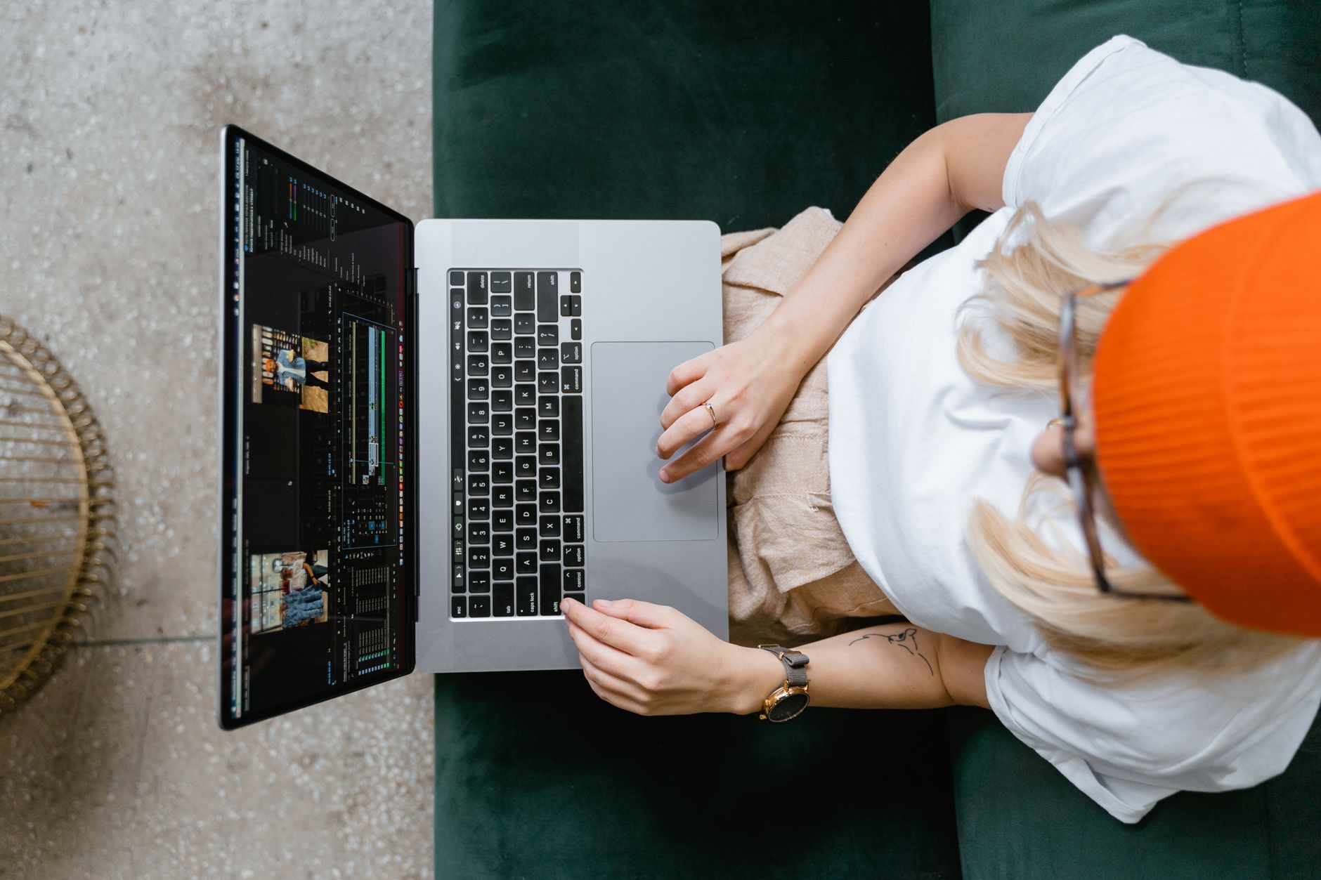 Top view of a woman in an orange beanie editing video footage on a laptop while sitting on a green sofa