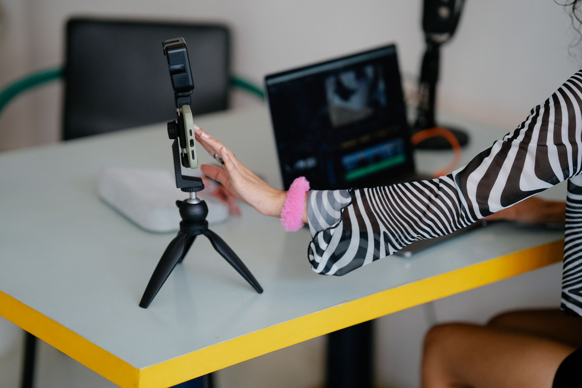 A woman adjusting a smartphone on a small tripod beside a laptop showing video editing software