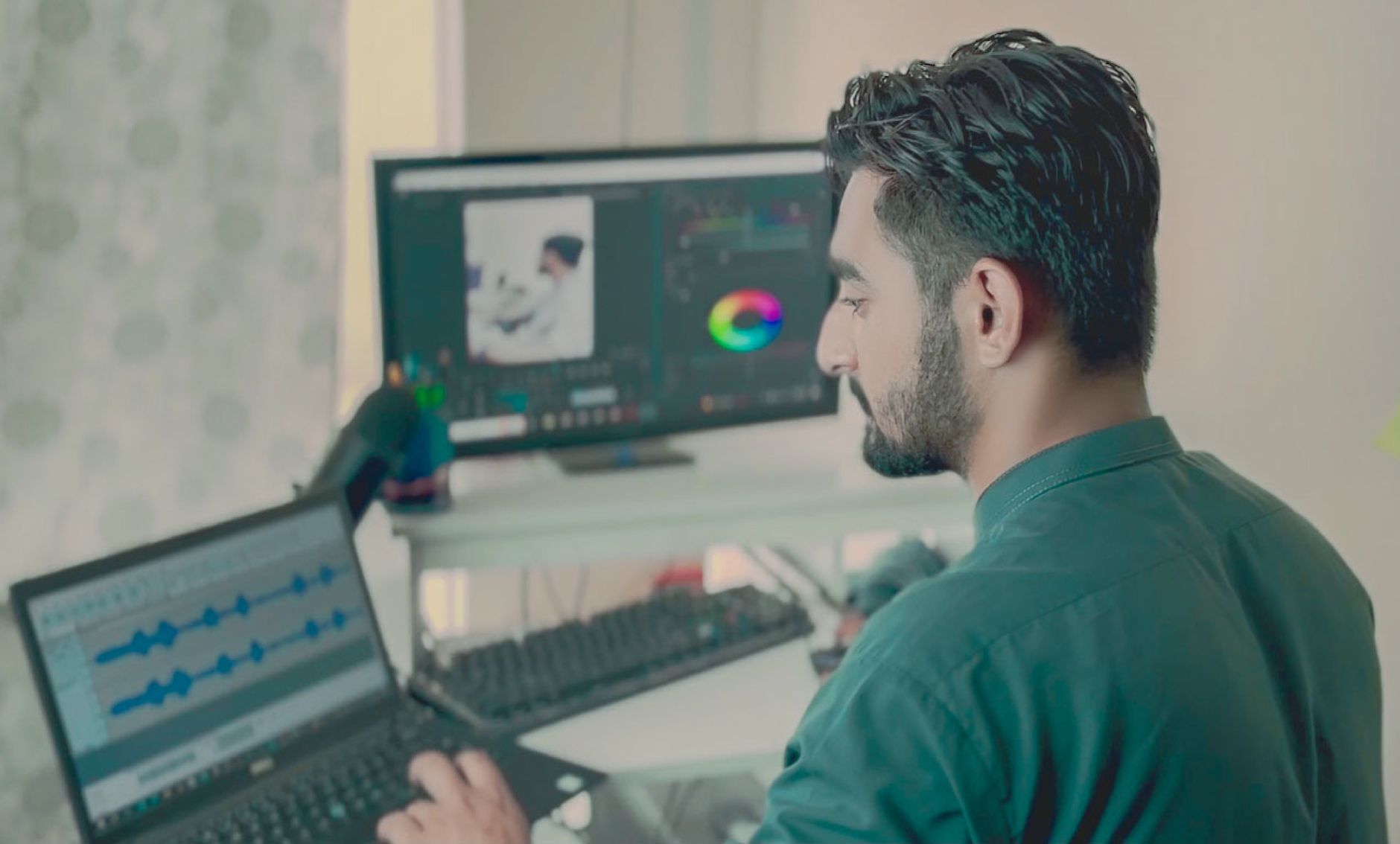 A man working at a desk with a laptop and two monitors showing audio and colour editing tools