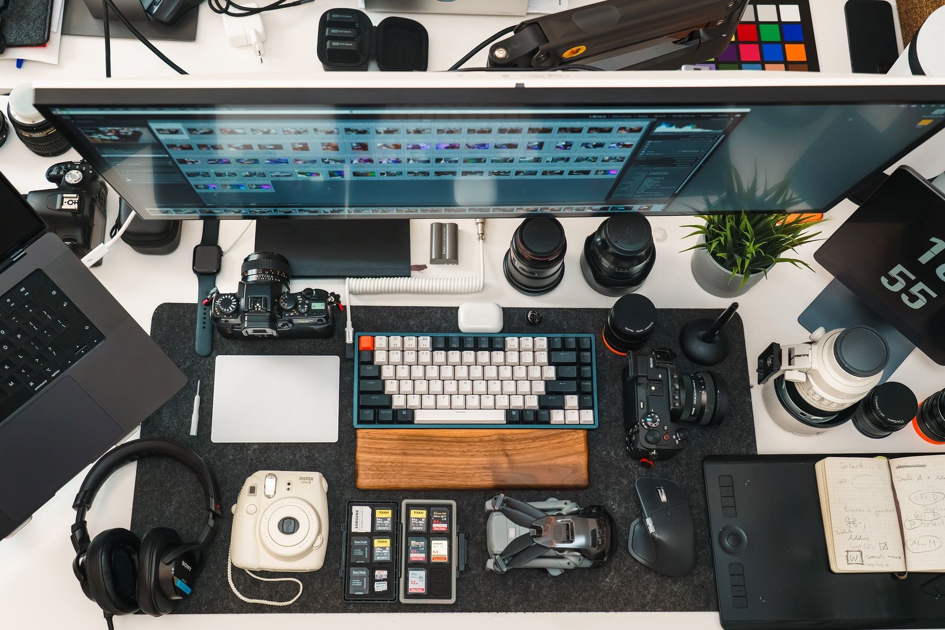 Flat lay of a fully equipped creator desk with cameras, lenses, a keyboard, headphones and a large monitor